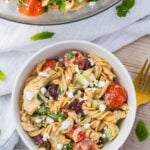 A bowl of gluten free pasta salad featuring rotini, cherry tomatoes, olives, cucumbers, and feta cheese on a textured cloth. A larger bowl of the same salad is partially visible above. A gold fork is placed beside the bowl. Parsley leaves are scattered around.