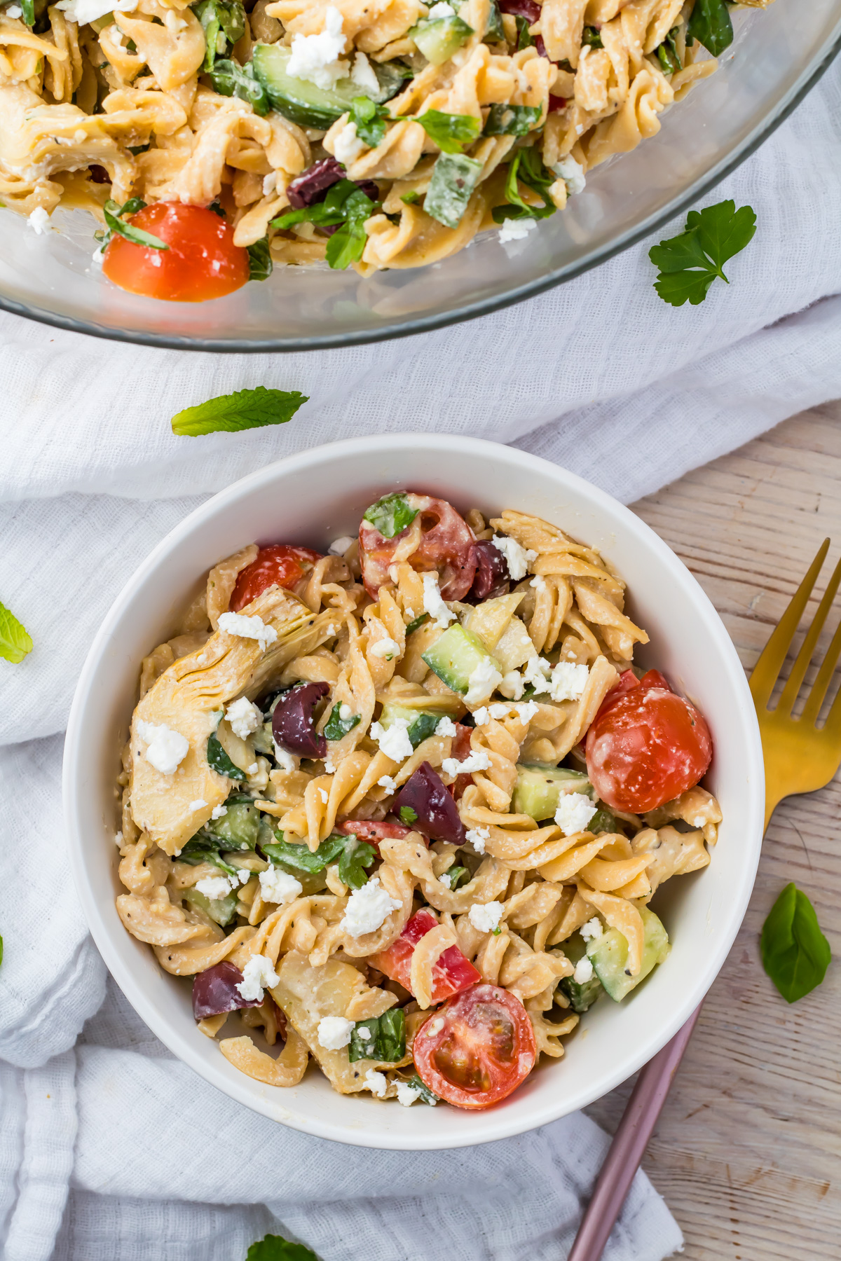 A bowl of gluten free pasta salad featuring rotini, cherry tomatoes, olives, cucumbers, and feta cheese on a textured cloth. A larger bowl of the same salad is partially visible above. A gold fork is placed beside the bowl. Parsley leaves are scattered around.