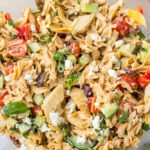 A close-up of a colorful pasta salad in a clear bowl. The salad features fusilli pasta, cherry tomatoes, cucumbers, feta cheese, olives, and parsley. A serving spoon is partially visible in the bowl.
