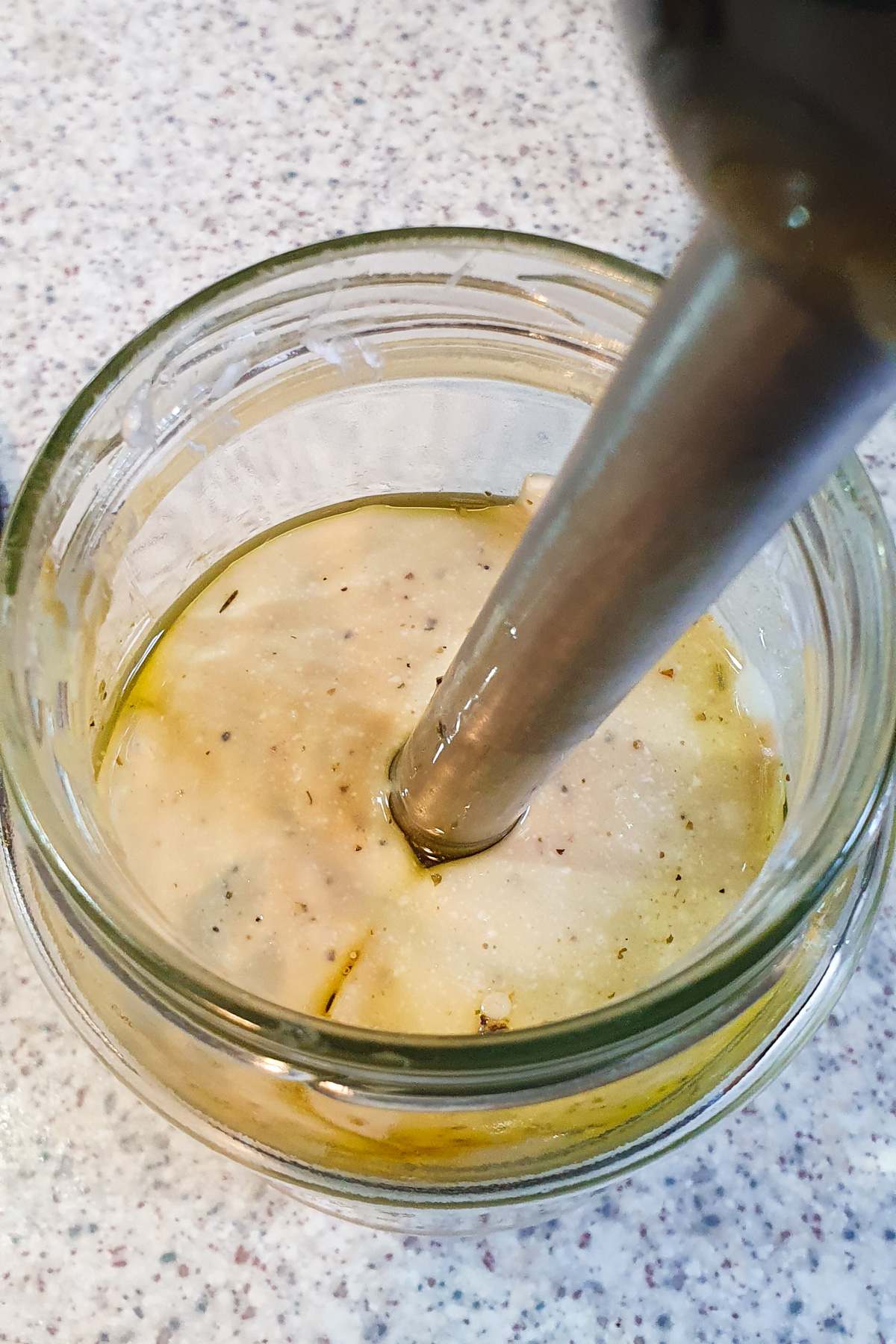A jar is filled with ingredients being blended by an immersion blender on a speckled countertop. The mixture appears creamy with visible oil and pepper.