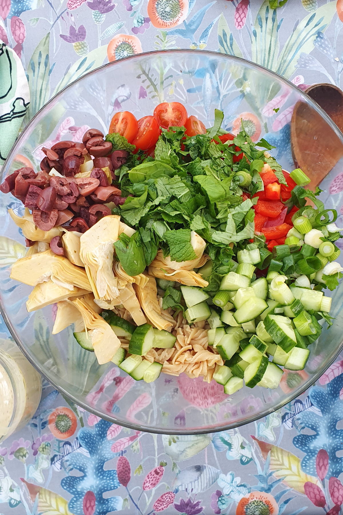 A glass bowl filled with chopped ingredients on a floral tablecloth. Visible are cherry tomatoes, sliced olives, red bell peppers, cucumbers, artichoke hearts, and fresh greens, ready to be mixed into a salad.