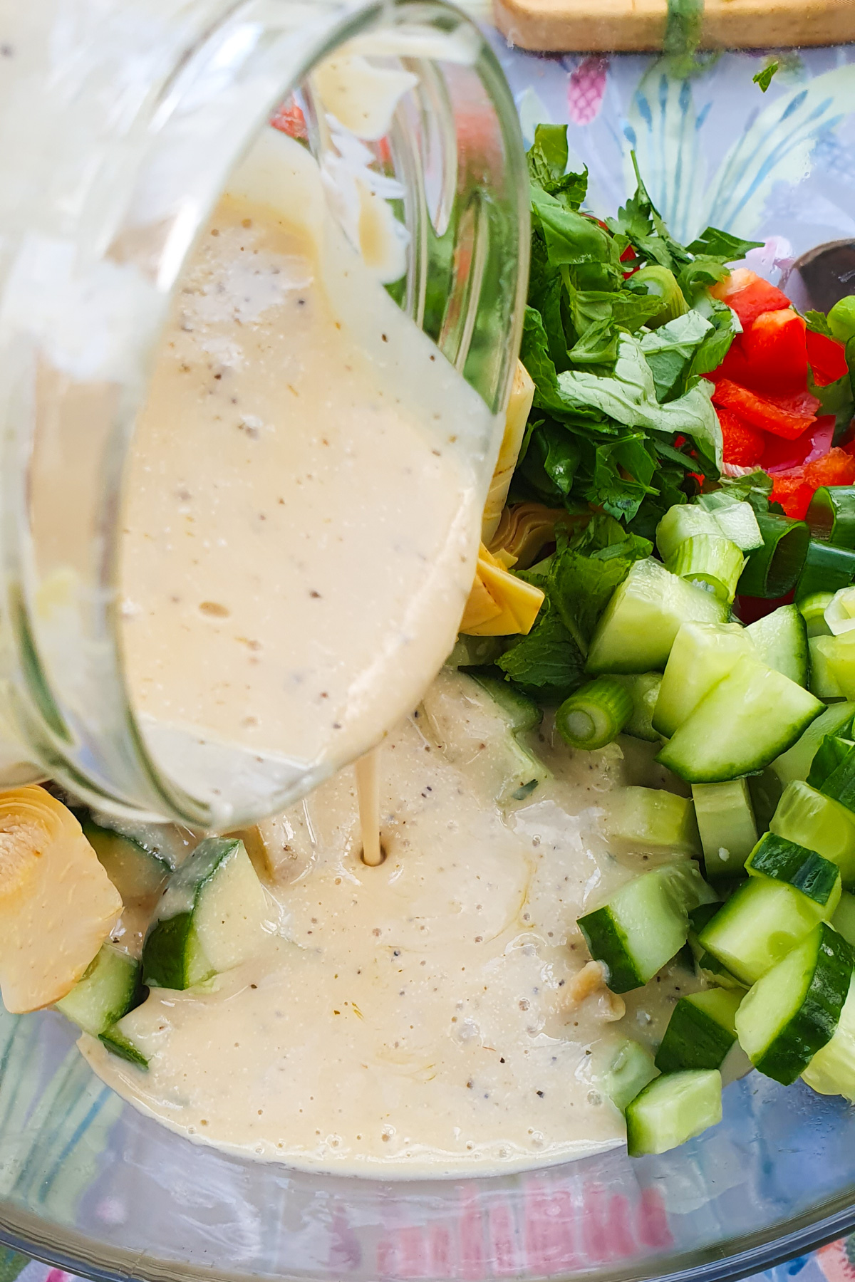 A creamy dressing is being poured from a jar onto a pasta salad with cucumbers, red bell peppers, leafy greens, and artichoke hearts in a bowl.