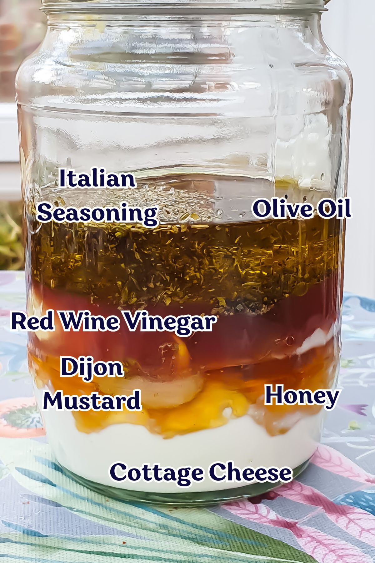 A glass jar filled with layers of ingredients labeled from top to bottom: Italian seasoning, olive oil, red wine vinegar, Dijon mustard, honey, and cottage cheese. The jar is on a floral-patterned surface.