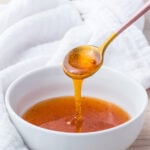 A spoon drizzling hot honey sauce into a white bowl, with a white cloth in the background on a light wooden surface.