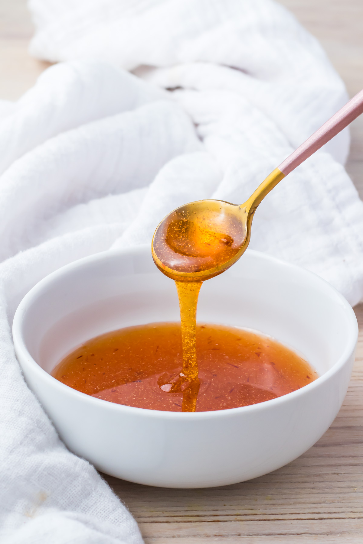 A spoon drizzling hot honey sauce into a white bowl, with a white cloth in the background on a light wooden surface.