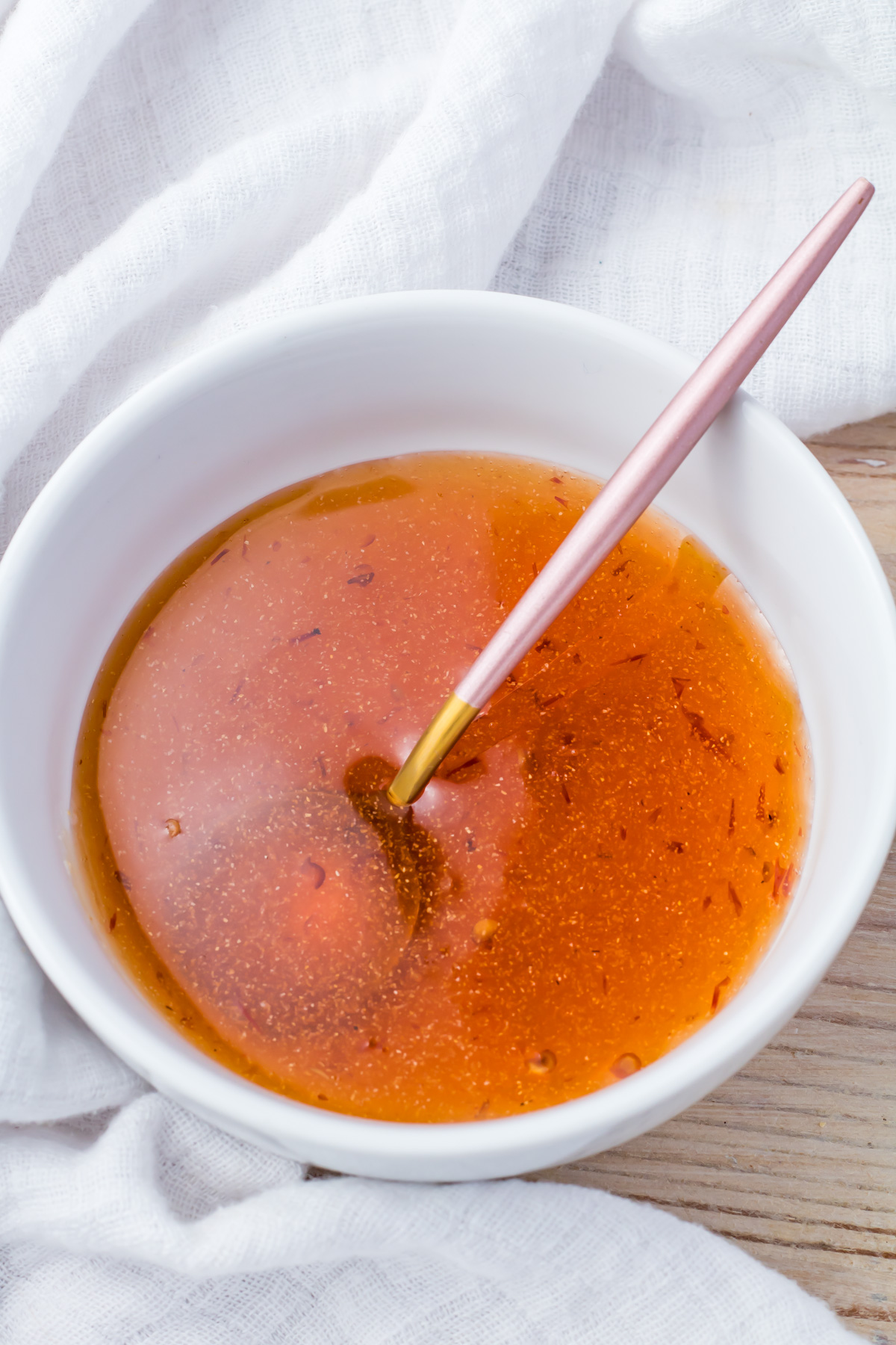 A white bowl filled with hot honey sauce. A pink-handled spoon is dipped into it. The bowl is placed on a light wooden surface with a white cloth partially visible.