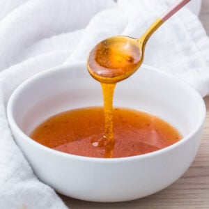 A golden spoon drizzling hot honey sauce into a white bowl, with a textured white cloth in the background on a wooden surface.