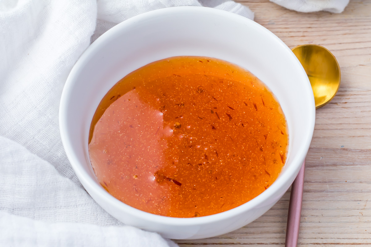 A white bowl filled with amber-colored hot honey sauce, placed on a wooden surface. A gold spoon is partially visible on the right, and a white fabric is draped nearby.