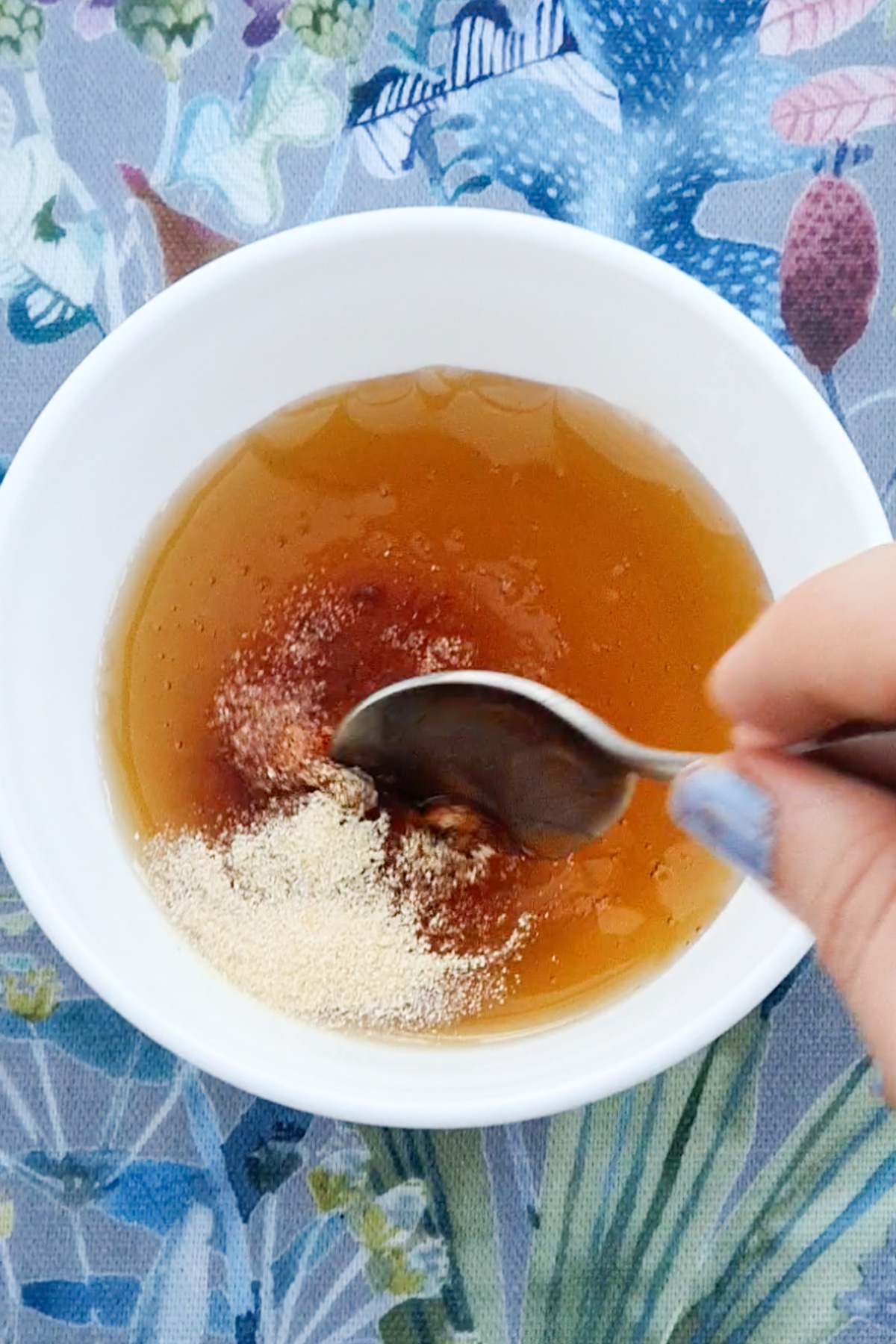 A bowl with honey, is on a floral-patterned tablecloth. A hand with painted nails is using a spoon to mix in a sprinkle of garlic powder and hot sauce.