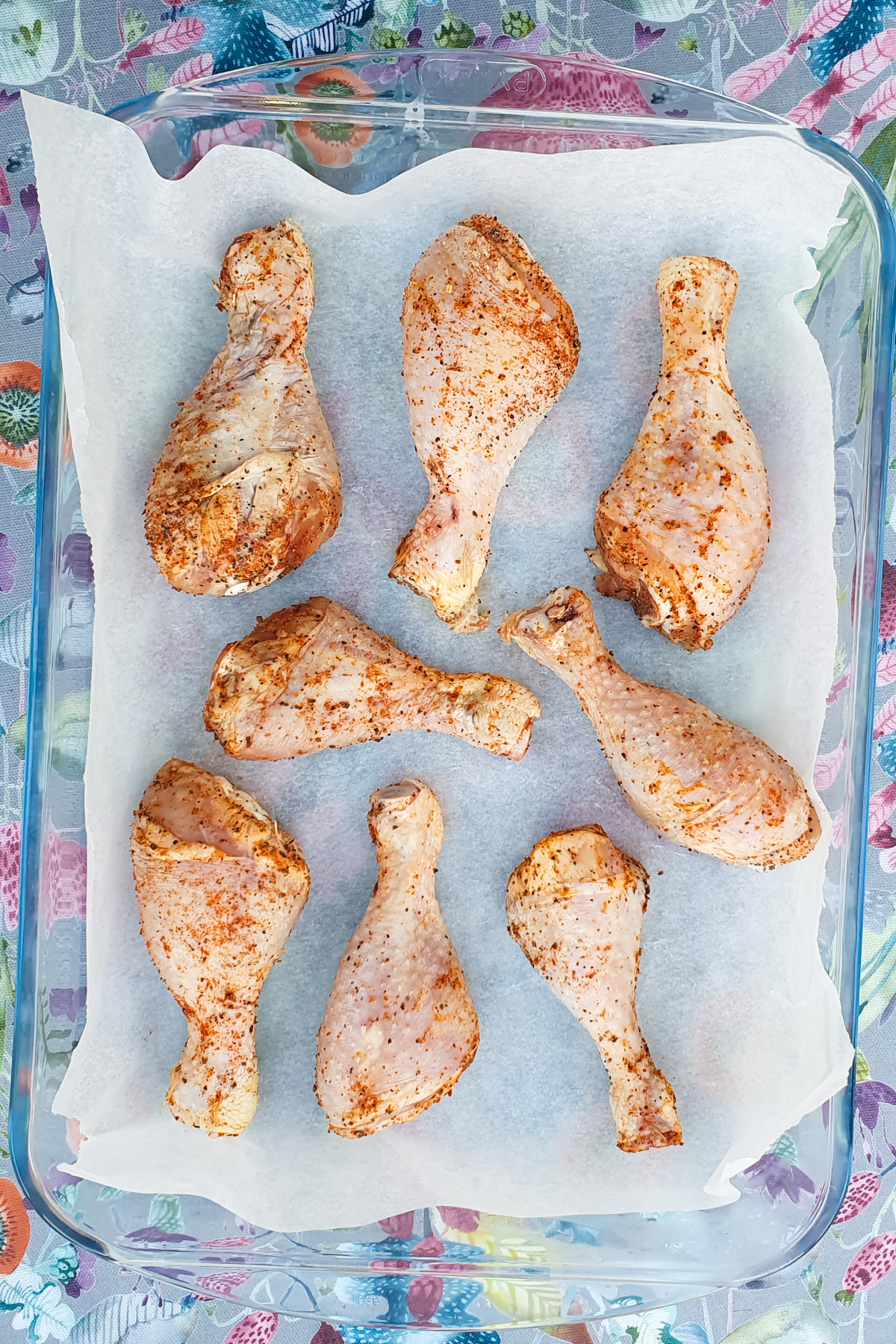 Eight seasoned chicken drumsticks are arranged on a parchment-lined baking tray. The seasoning includes spices that give the chicken a reddish-orange tint. The tray is set against a colorful, patterned background.