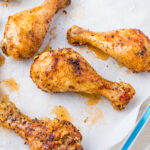 Five golden-brown oven baked chicken drumsticks on a parchment-lined glass baking dish. The drumsticks are seasoned with herbs and spices, with a few oil drips around them.
