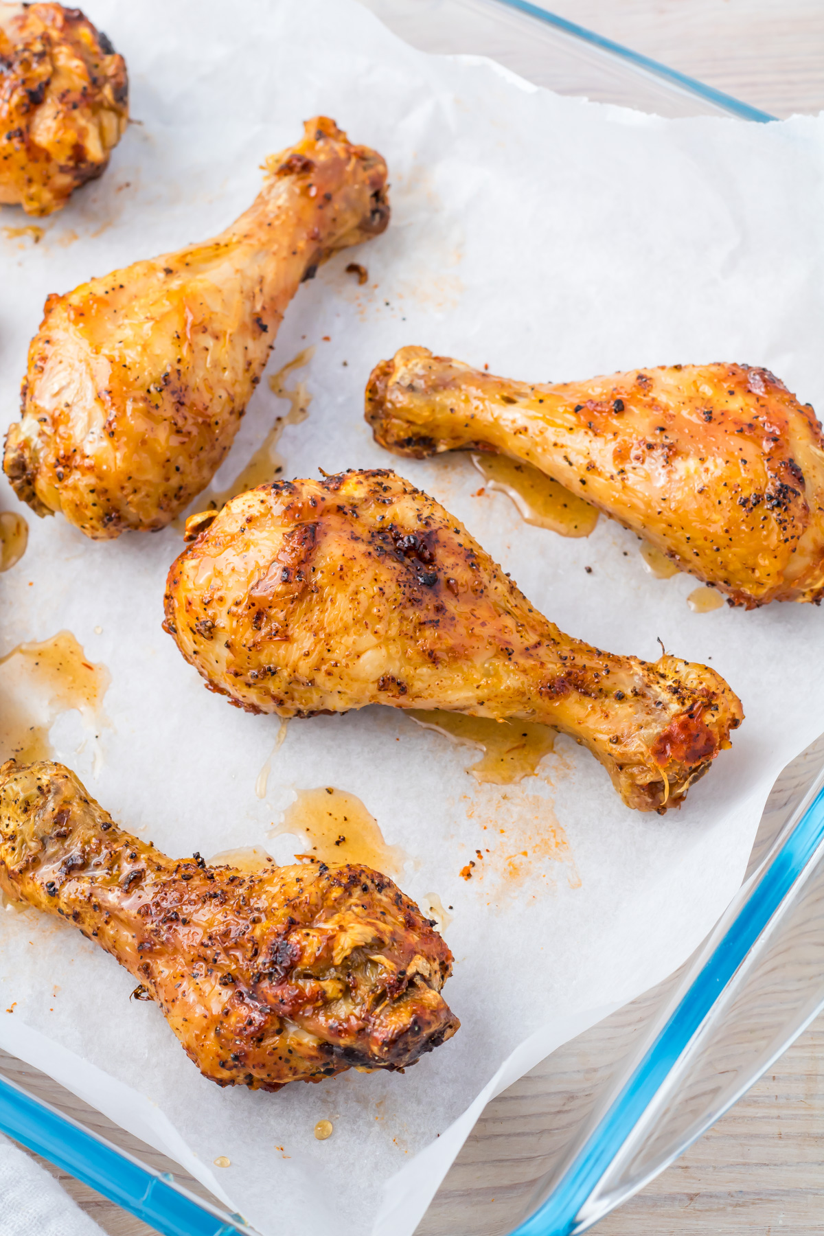 Five golden-brown oven baked chicken drumsticks on a parchment-lined glass baking dish. The drumsticks are seasoned with herbs and spices, with a few oil drips around them.