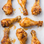 Eight crispy, golden-brown roasted chicken drumsticks are arranged on a sheet of white parchment paper. The chicken is seasoned with spices, and some juices glisten on the surface, enhancing their appetizing appearance.