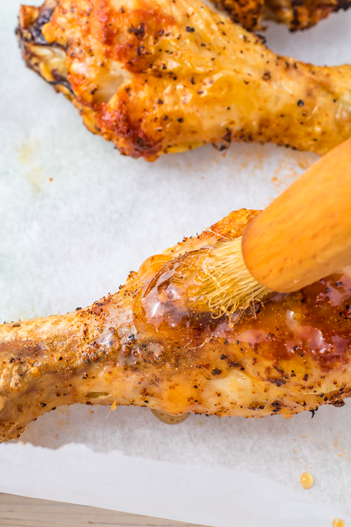 Close-up of a seasoned and roasted chicken drumstick being brushed with hot honey sauce. The chicken appears golden brown with visible spices, and the brush has a wooden handle. The background is white parchment.