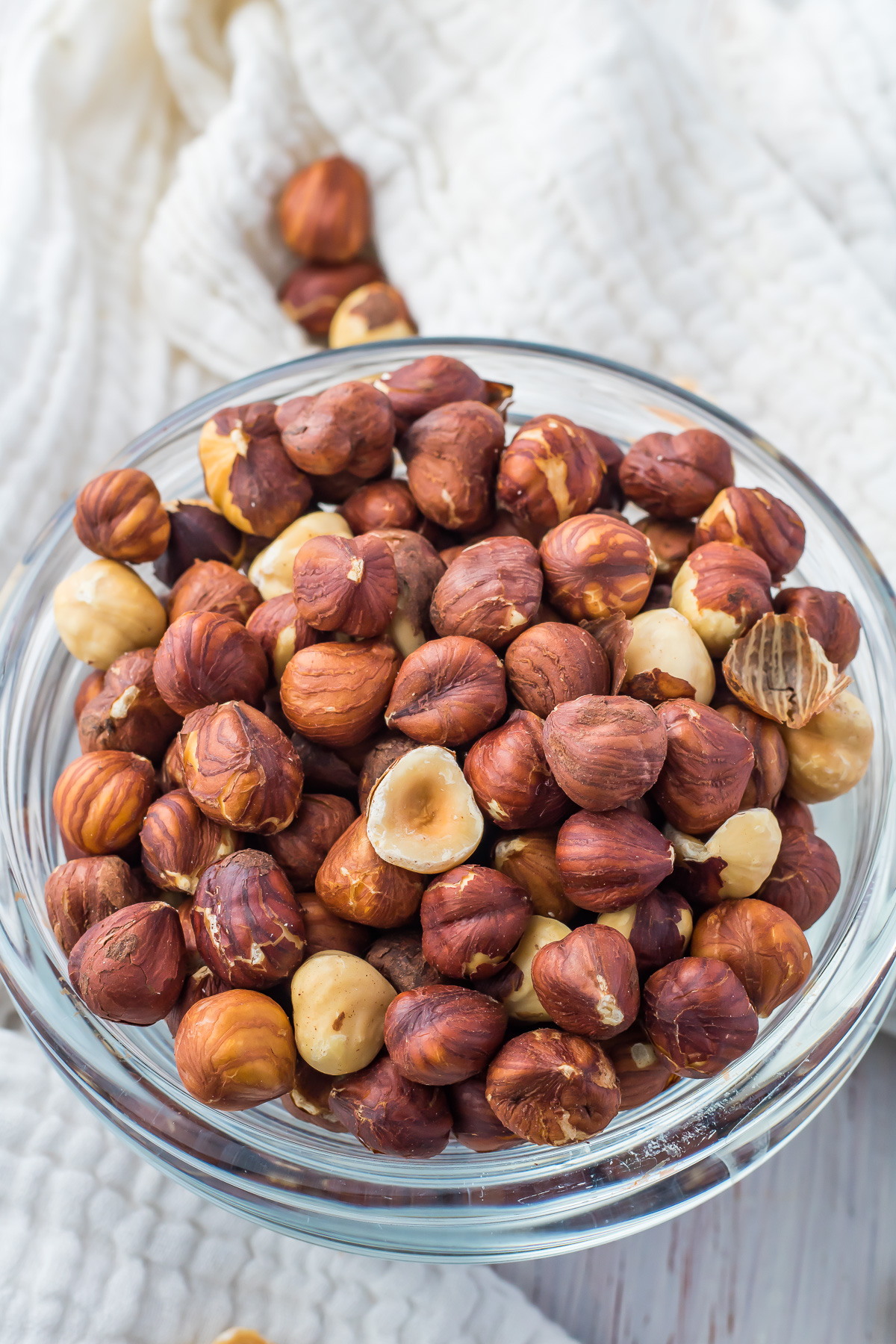 A glass bowl filled with whole air fryer hazelnuts, some with skins partially removed, placed on a textured white cloth background.