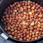 A large batch of hazelnuts with shells on is piled inside the basket of an air fryer, ready for roasting. The air fryer is black and has a round shape. The hazelnuts have a rich brown color with various shades.