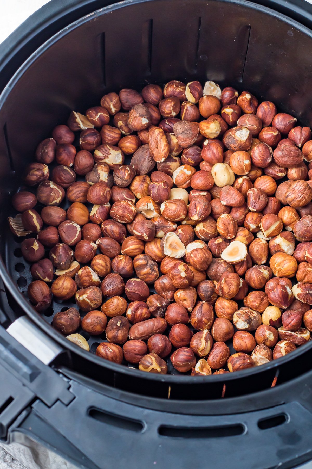 A large batch of hazelnuts with shells on is piled inside the basket of an air fryer, ready for roasting. The air fryer is black and has a round shape. The hazelnuts have a rich brown color with various shades.
