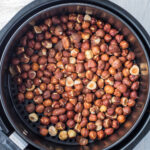 A black air fryer basket filled with roasted hazelnuts. The nuts have a rich brown color, and some shells are cracked. A textured white cloth is partially visible in the background on a light surface.