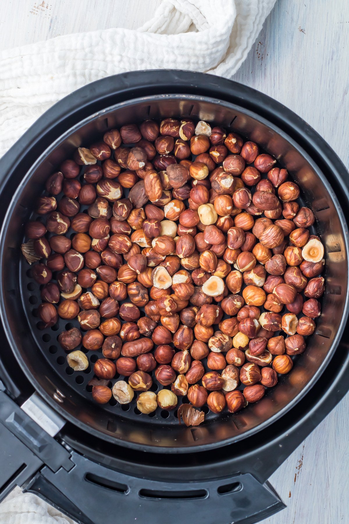 A black air fryer basket filled with roasted hazelnuts. The nuts have a rich brown color, and some shells are cracked. A textured white cloth is partially visible in the background on a light surface.