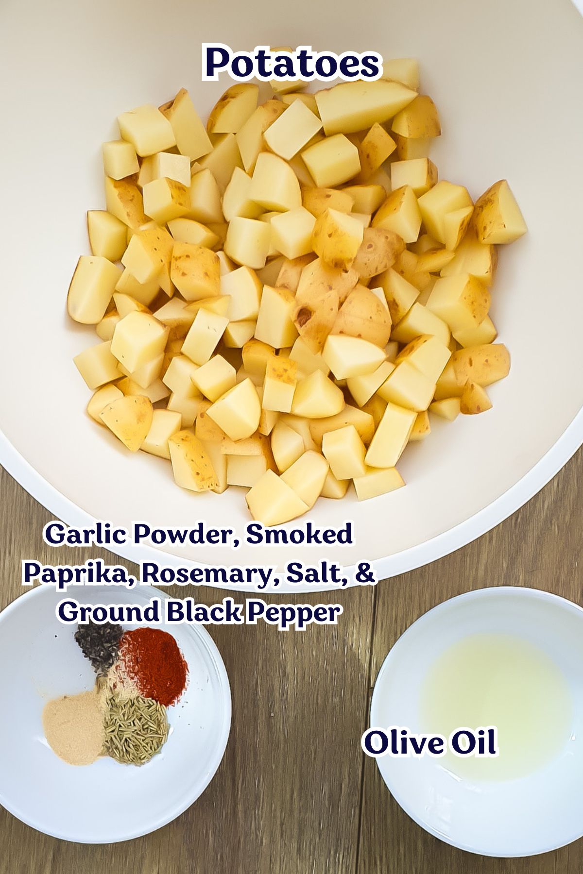 A bowl of diced potatoes, a small bowl of olive oil, and a plate with garlic powder, smoked paprika, rosemary, salt, and ground black pepper on a wooden surface.