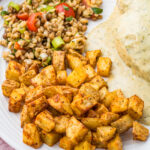 A plate with air fryer diced potatoes, a serving of grain salad with tomatoes and herbs, and a portion of chicken covered in creamy white sauce.