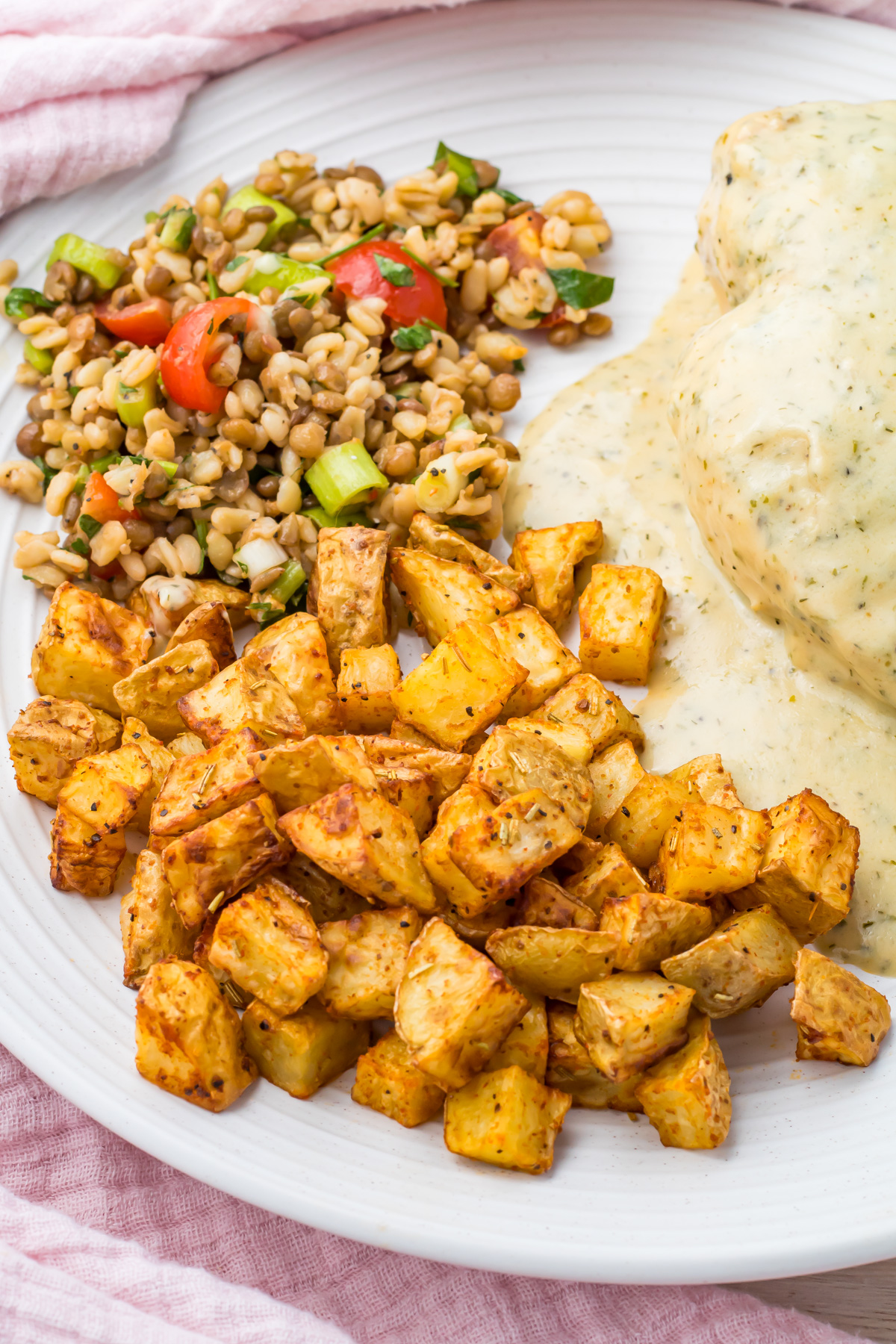 A plate with air fryer diced potatoes, a serving of grain salad with tomatoes and herbs, and a portion of chicken covered in creamy white sauce.