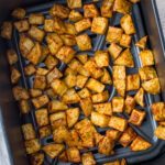 Seasoned diced potatoes are spread evenly on a black baking tray, showing a golden-brown exterior.