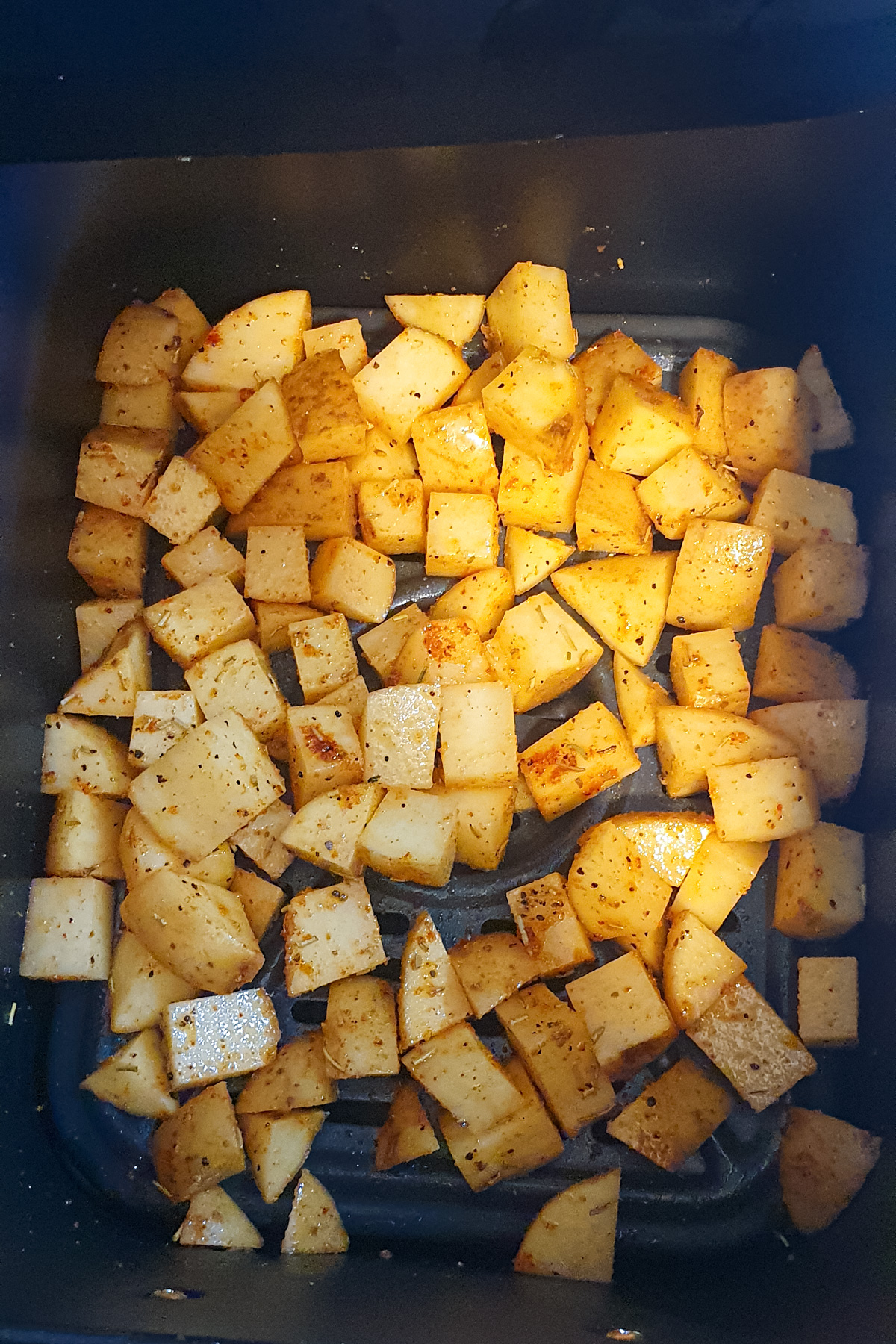 Diced potatoes coated with seasoning are spread evenly inside an air fryer basket, ready to be cooked.