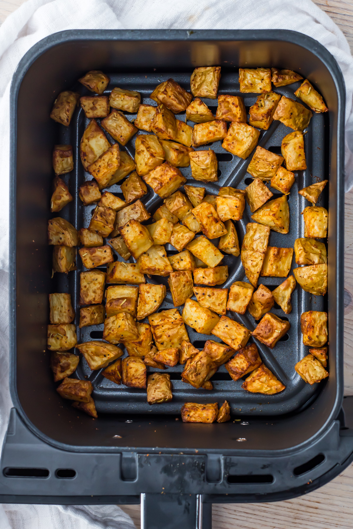 Cubed potatoes seasoned and cooked in an air fryer basket, placed on a light-colored cloth on a wooden surface.