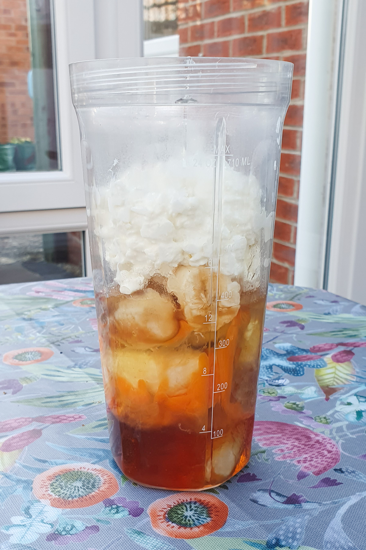 A clear blender cup containing ice, chopped bananas, honey, and a layer of yogurt, placed on a colorful tablecloth.
