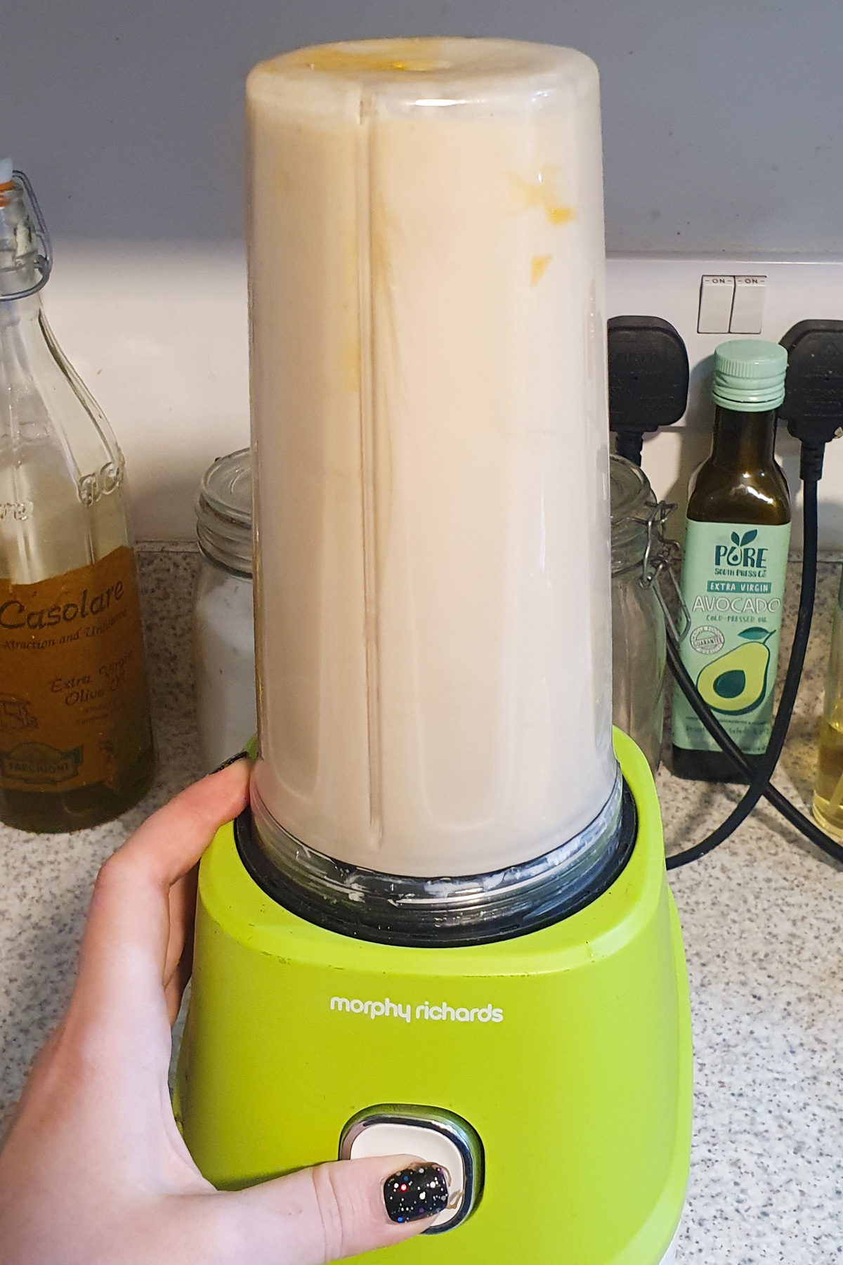 A hand operates a green Morphy Richards blender filled with a light-colored mixture on a kitchen counter surrounded by various bottles of oil.