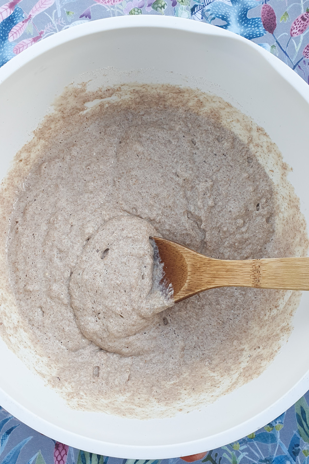 A white mixing bowl containing a thick, light brown batter being stirred with a wooden spoon, placed on a floral-patterned surface.