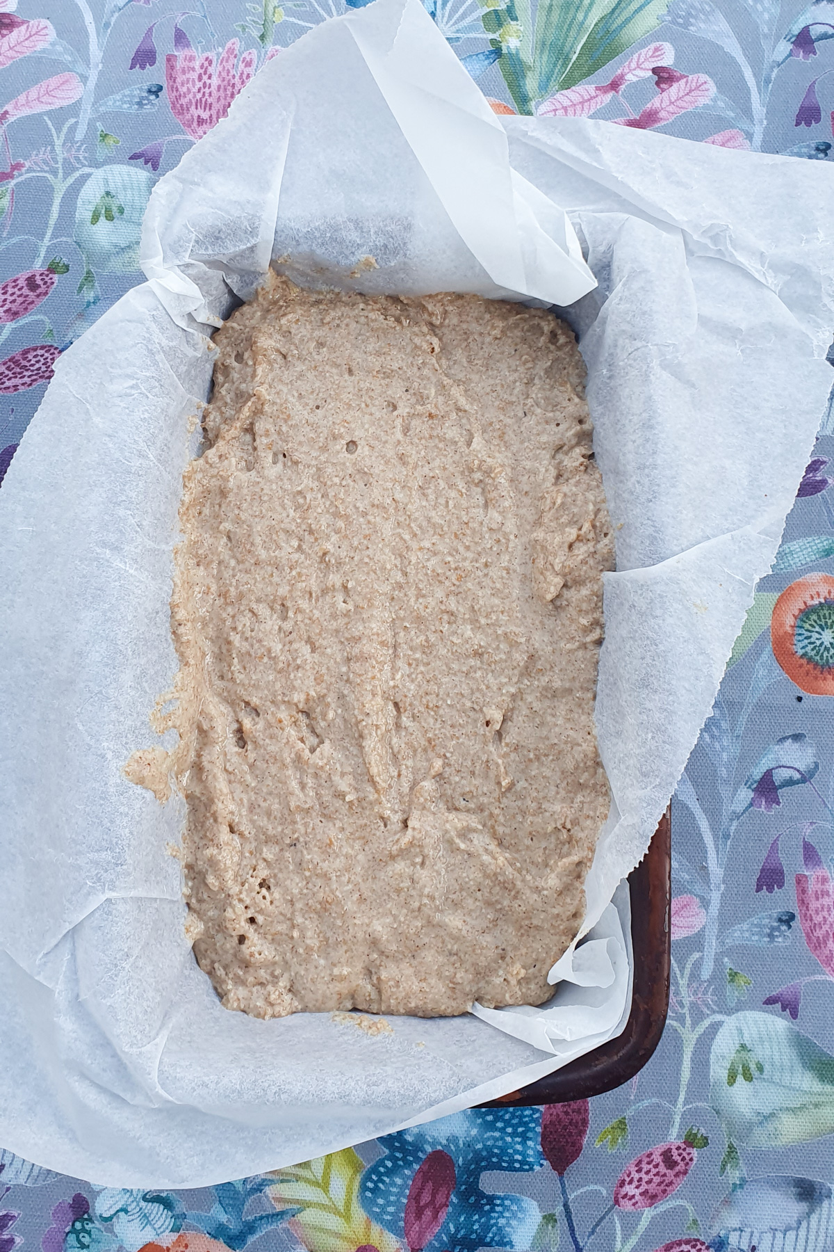 A rectangular baking tin lined with parchment paper is filled with unbaked banana bread dough, resting on a colorful floral-patterned surface.