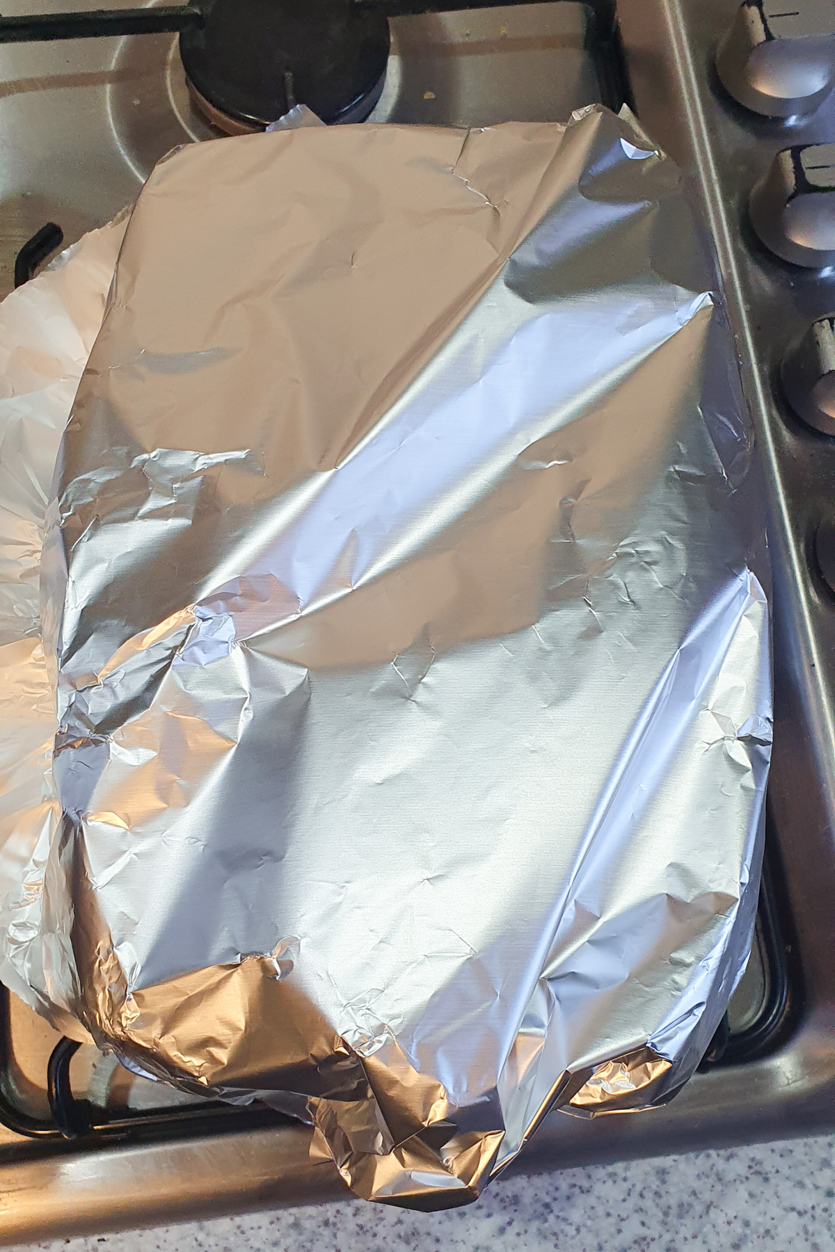 A baking tray covered with aluminum foil sits on a gas stove next to the burner controls.