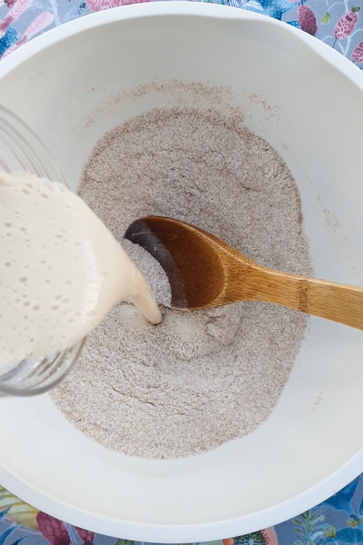 Batter is being poured from a glass container into a white bowl of dry ingredients, with a wooden spoon resting in the bowl.