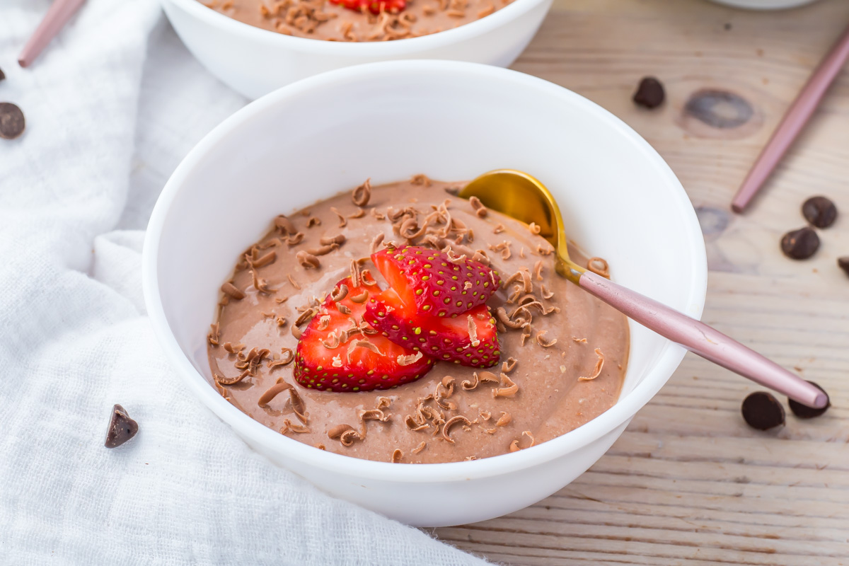 A white bowl filled with chocolate cottage cheese mousse, topped with sliced strawberries and chocolate shavings, with a gold spoon on the side.