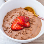 A white bowl filled with chocolate cottage cheese mousse, topped with sliced strawberries and chocolate shavings, with a gold spoon on the side.
