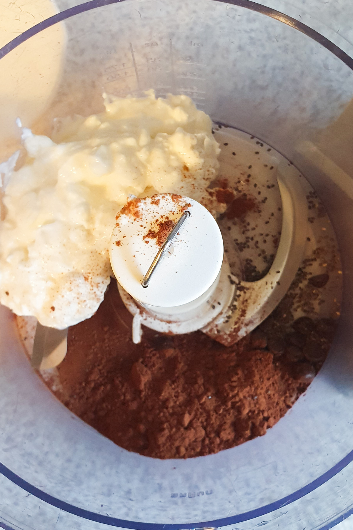 A food processor bowl containing cocoa powder, cottage cheese, chia seeds, and a sprinkle of coffee powder, ready to be blended.