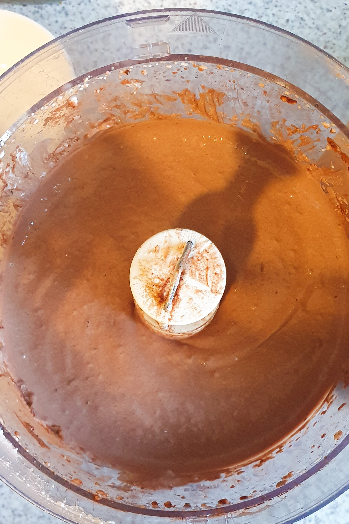 A food processor bowl filled with smooth chocolate batter, with some batter on the lid and sides.