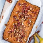A loaf of healthy banana bread topped with pecans sits in a parchment-lined baking pan, with a serrated knife, pecans, and bananas nearby on a white cloth.