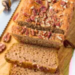 Sliced healthy banana bread topped with chopped pecans on a wooden cutting board, with a serrated knife and two bananas in the background.