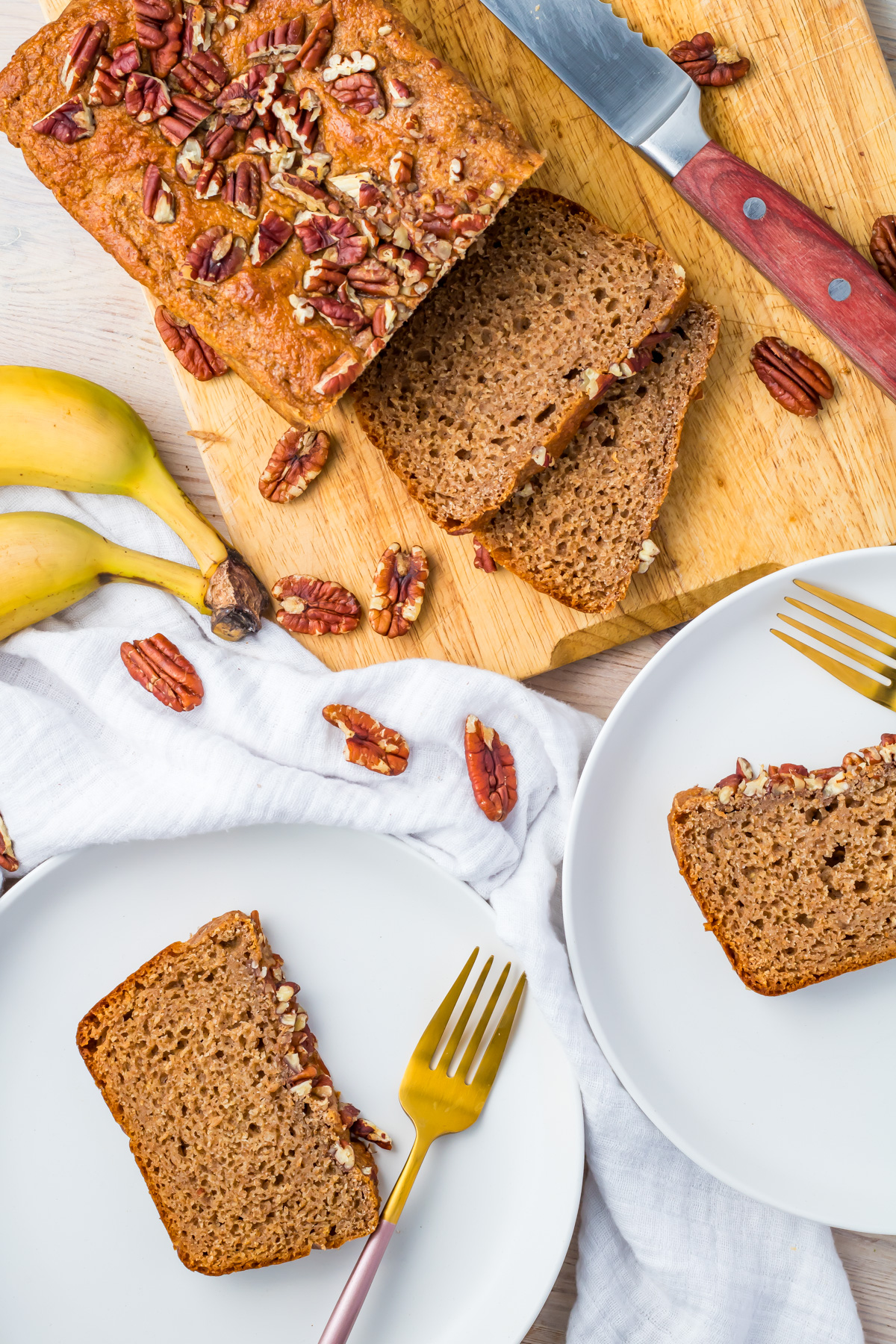 A loaf of healthy banana bread topped with pecans is sliced on a wooden board, with two plated slices and gold forks beside scattered pecans and bananas.