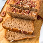 Sliced loaf of healthy banana bread topped with chopped pecans on a wooden cutting board, with a few pecans scattered nearby.