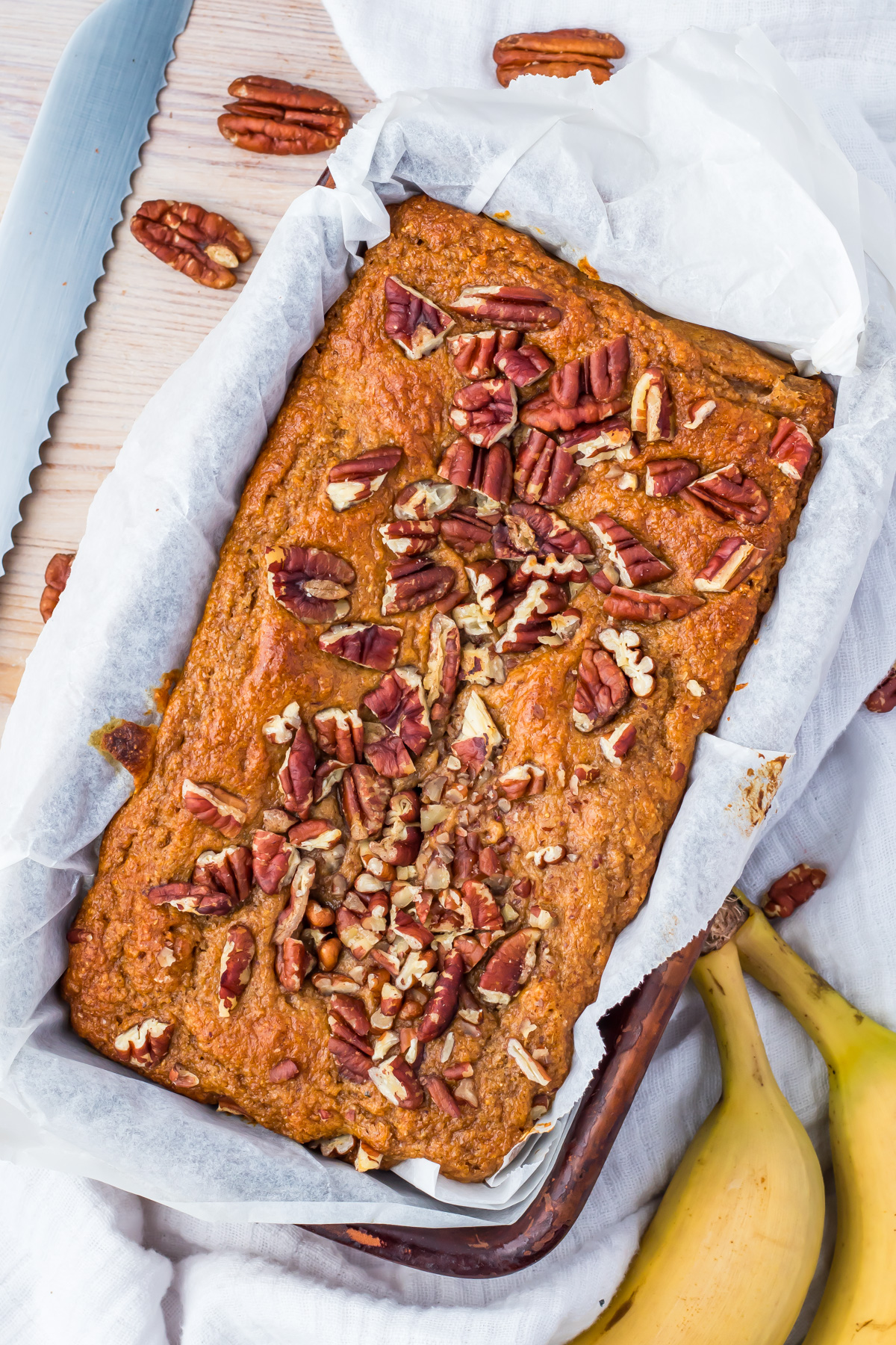 A loaf of healthy banana bread topped with pecans sits in a parchment-lined baking pan, with a serrated knife, pecans, and bananas nearby on a white cloth.