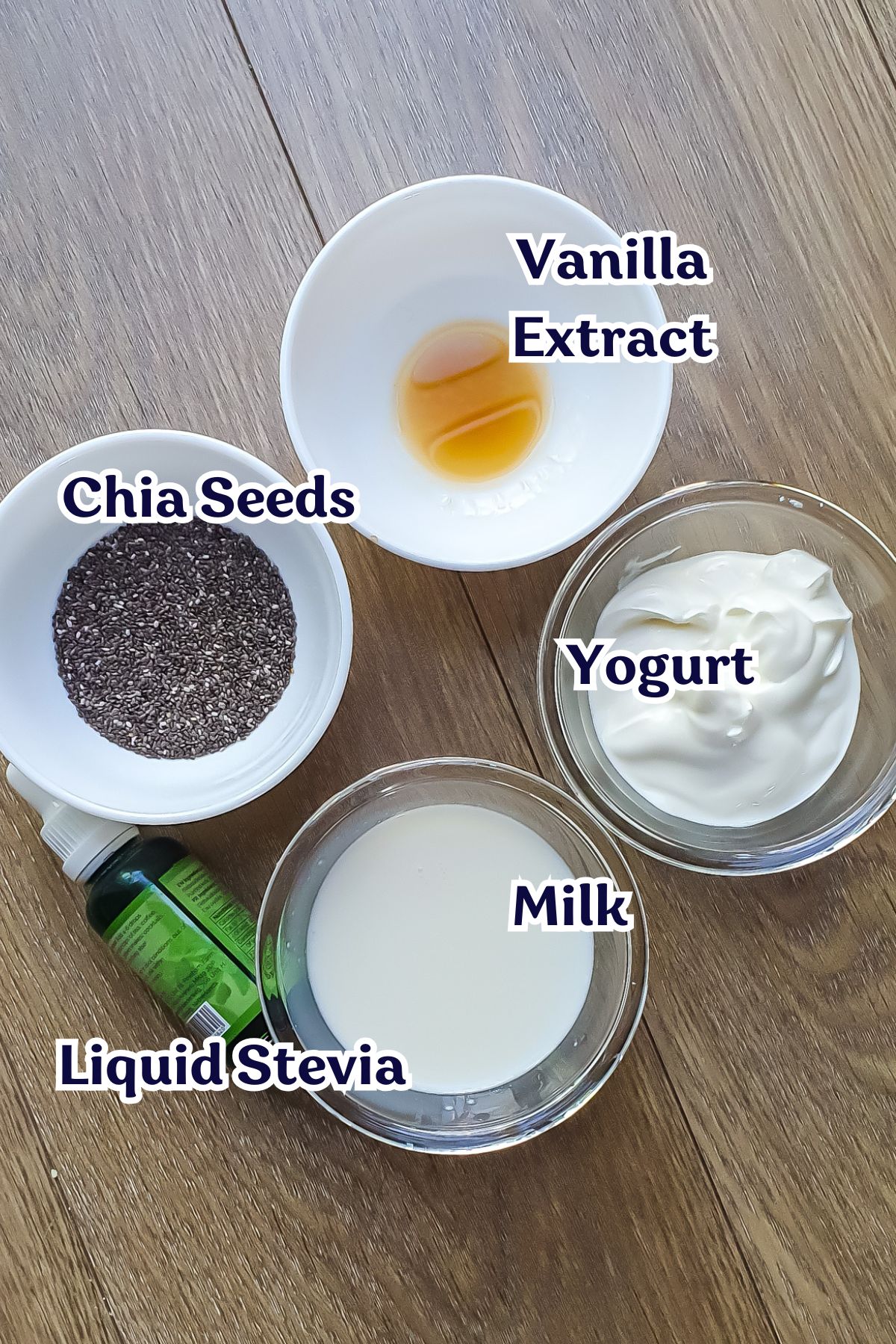 Five labeled ingredients in separate bowls or containers on a wooden surface: chia seeds, vanilla extract, yogurt, milk, and liquid stevia.