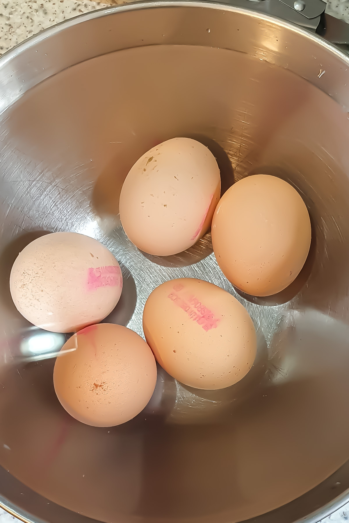 Five brown air fryer hard boiled eggs with pink stamps are placed in ice water in a stainless steel mixing bowl on a kitchen countertop.