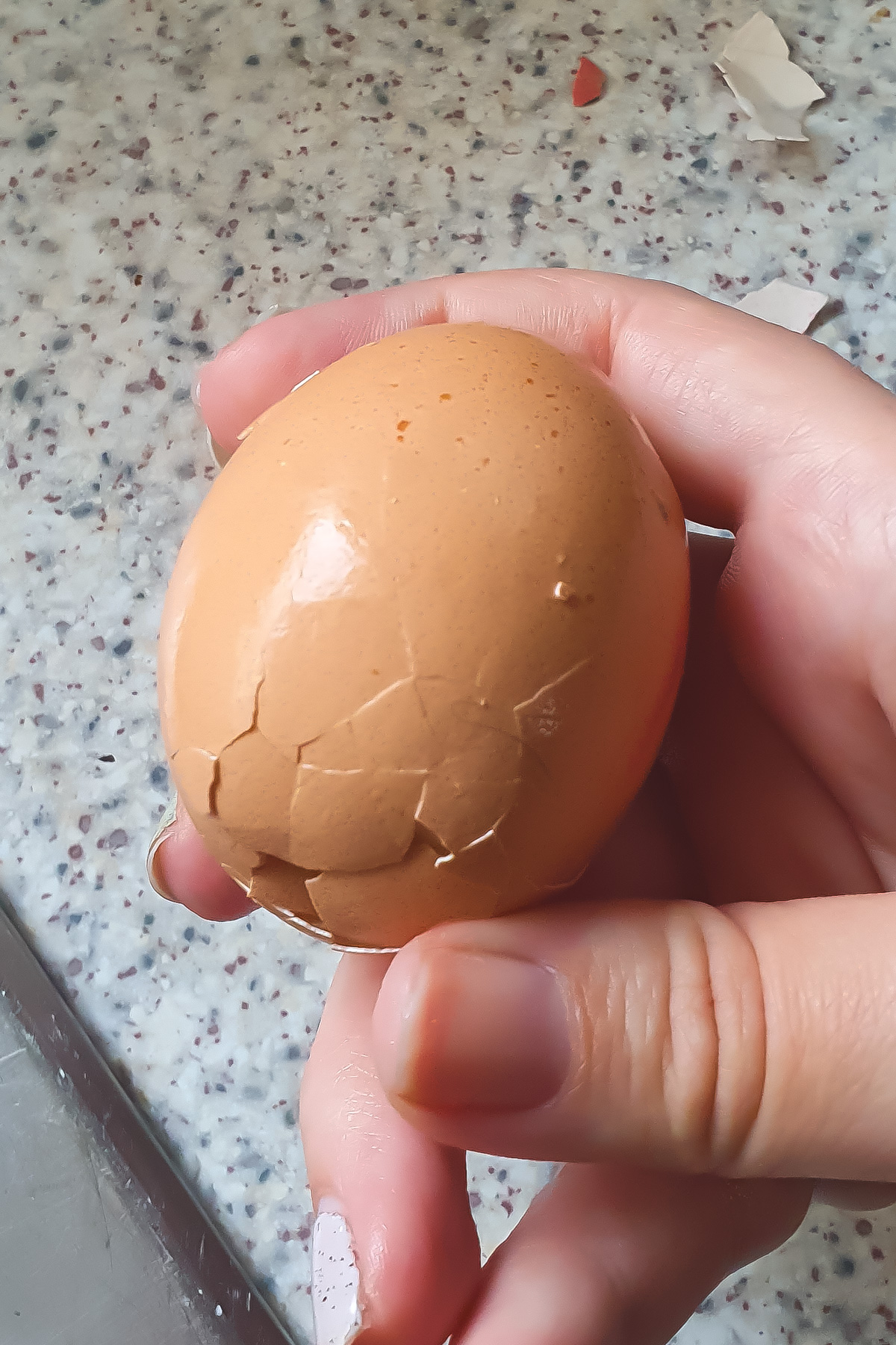 A hand holding a brown hard boiled egg with a cracked shell over a speckled countertop.