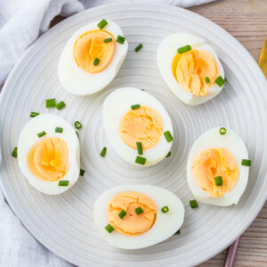 A white plate with six halved air fryer hard boiled eggs, each topped with chopped green chives, on a light wooden surface.