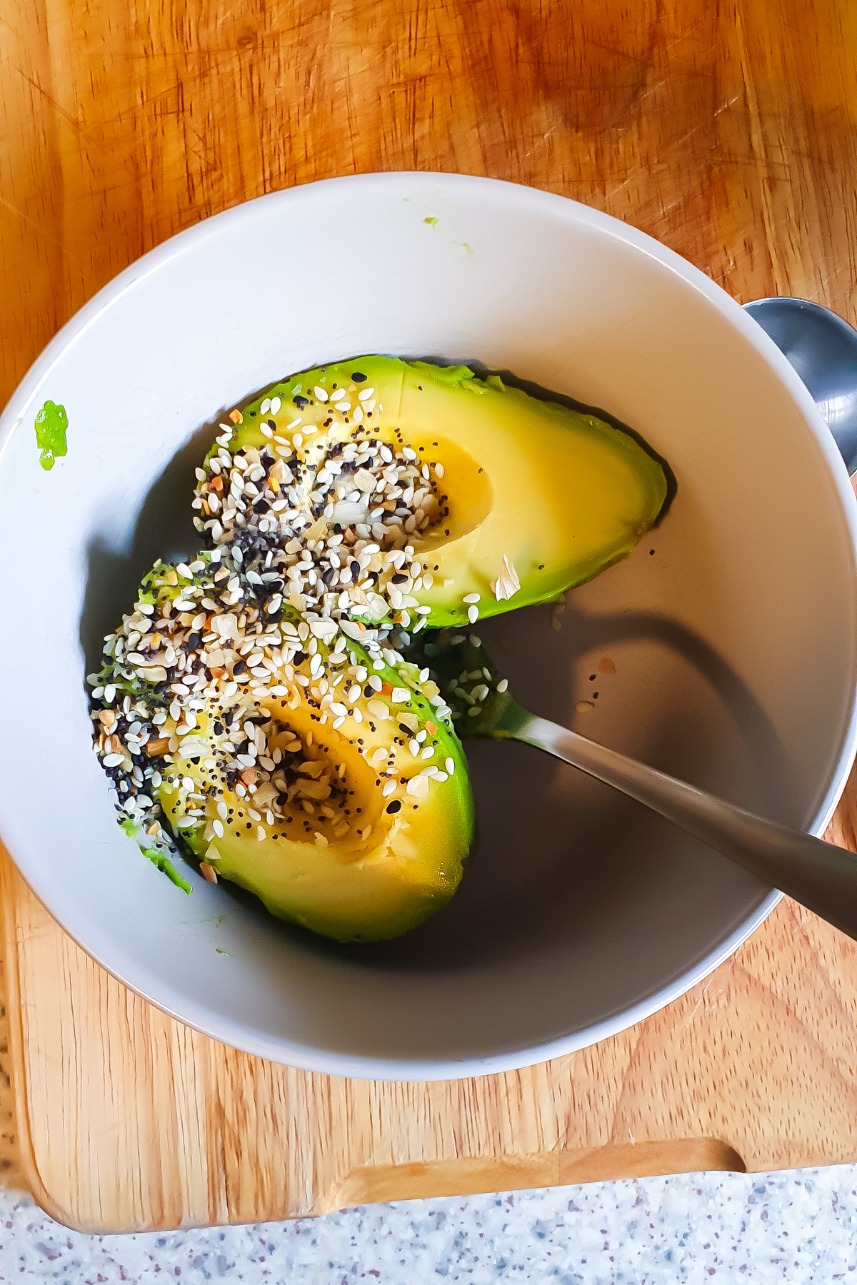 A halved avocado in a white bowl, topped with a seed seasoning mix, with a spoon beside it, on a wooden cutting board.