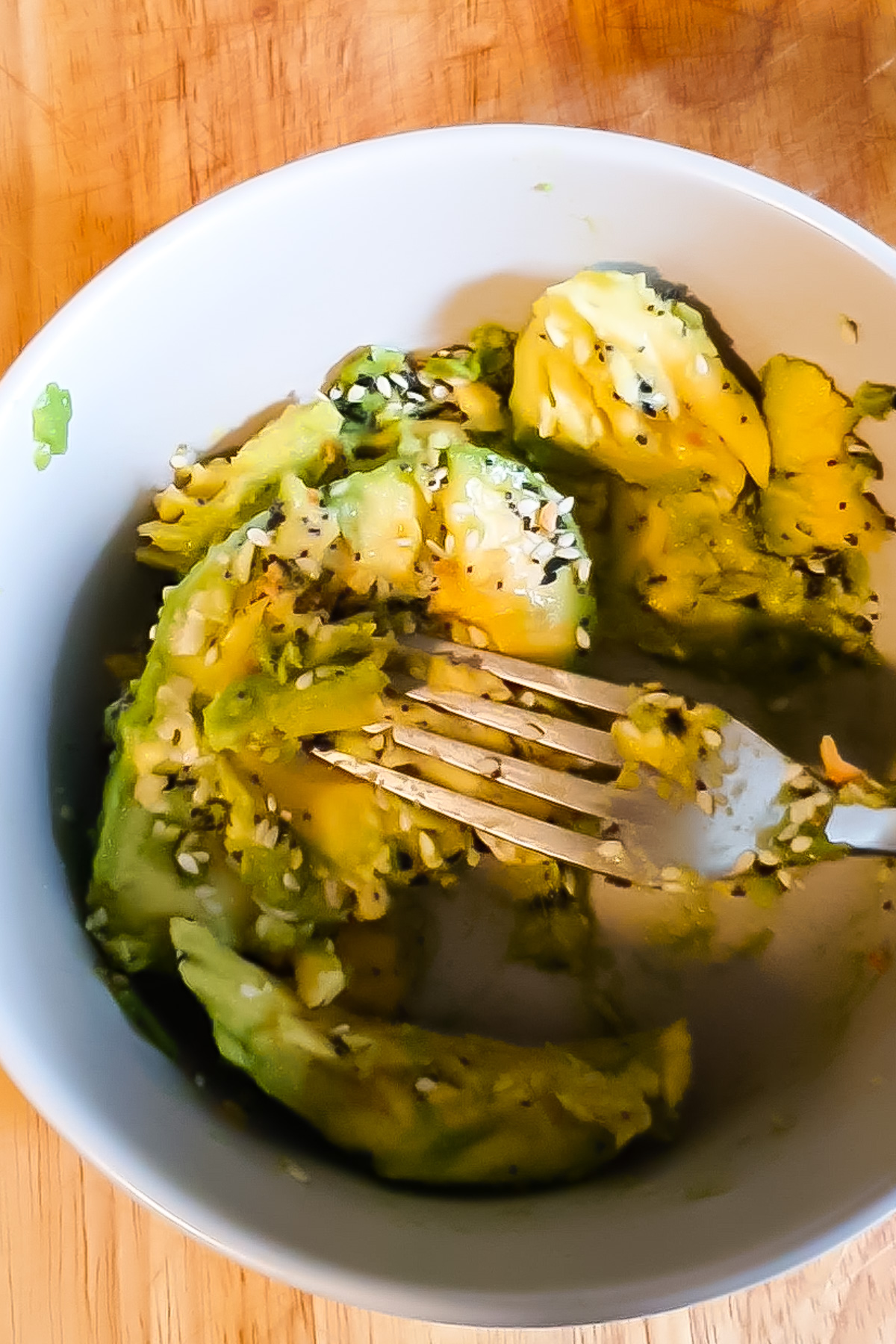 A halved avocado in a white bowl, topped with a seed seasoning mix, being mashed with a fork, on a wooden cutting board.